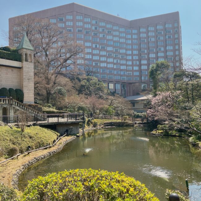 ホテル椿山荘東京の広大な日本庭園の景色。手前には池があり、その奥には緑豊かな木々が生い茂り、さらにその先にホテルの建物がそびえ立っている。庭園内には小川や橋、石造りの塔のような構造物も見え、自然と調和した景観が広がっている。