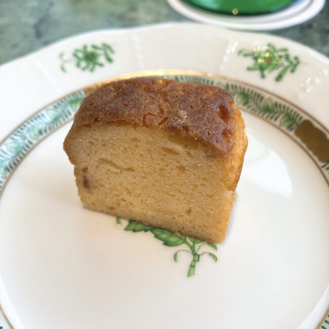 緑がかったマーブル模様のテーブルの上に置かれた、白い皿に載った一切れのパウンドケーキ。パウンドケーキは長方形で、表面は黄金色に焼き上げられ、上部にはわずかな割れ目が見える。内部はきめ細やかな黄色がかった生地。これぞホテルメイドのパウンドケーキ。