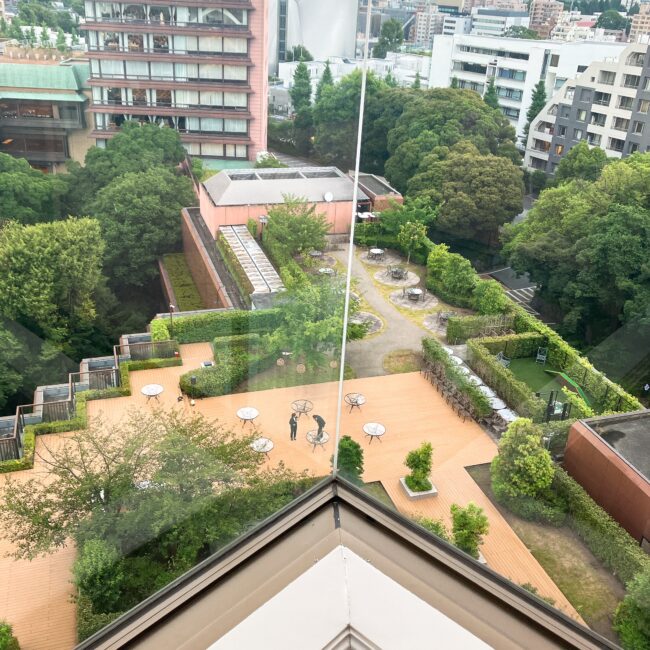 ホテル椿山荘東京パゴダルームの窓から見下ろす、5階テラスとドッグランの様子。ウッドデッキのテラスにはテーブルと椅子が配置され、緑豊かな庭園の中にドッグランの囲いが見える。当日は犬の姿はなかった。