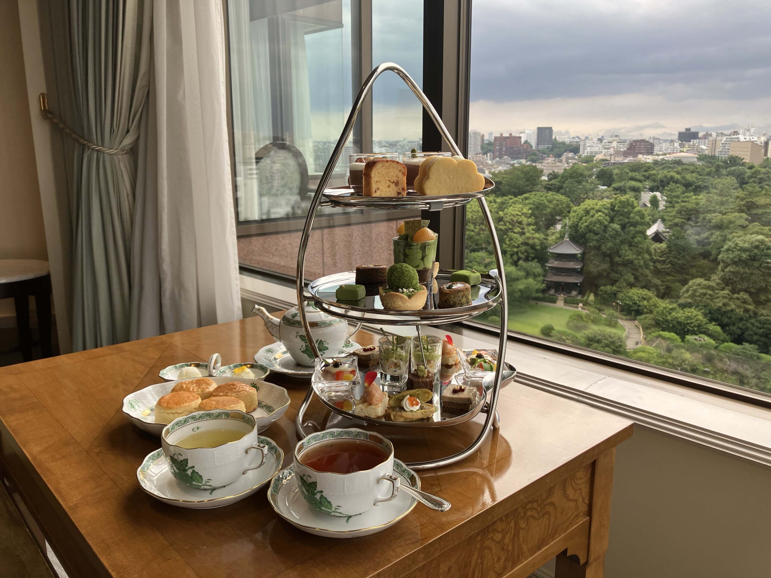 ホテル椿山荘東京のパゴダルームで提供されるプレミアムアフタヌーンティー。三段重ねのティースタンドには、彩り豊かなスイーツとセイボリーが並び、手前にはヘレンドのアポニーグリーンと思われるティーカップに入った紅茶が置かれている。窓の外には、緑豊かな日本庭園と、その向こうに広がる東京都心のビル群が見渡せる。