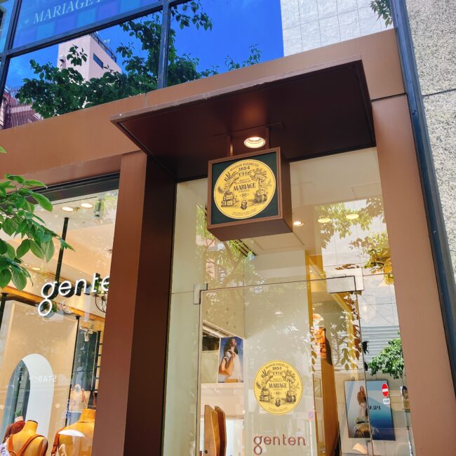 銀座松屋通りに面した建物の入り口付近。左手には「genten」と書かれたブティックのショーウィンドウが見える。その右隣に、円形の金色のマリアージュフレールのロゴ看板が掲げられている。入り口はガラス扉で、扉や壁には小さなマリアージュフレールのロゴシールも貼られている。画面中央右には、上へ続く階段の一部が見える。これがマリアージュフレール銀座松屋通り店(三和ビル2階)への推奨される入り口。
