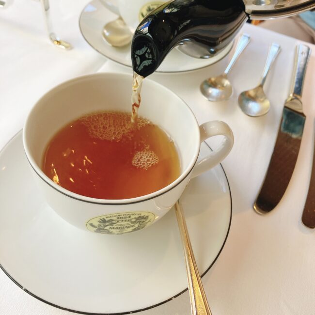 ティーポット「アール・デコ」から、紅茶が白いカップ&ソーサーに注がれている様子。カップにはマリアージュフレールのロゴが入っている。テーブルの上には白いクロスが敷かれ、シルバーのスプーンやナイフが並んでいる。