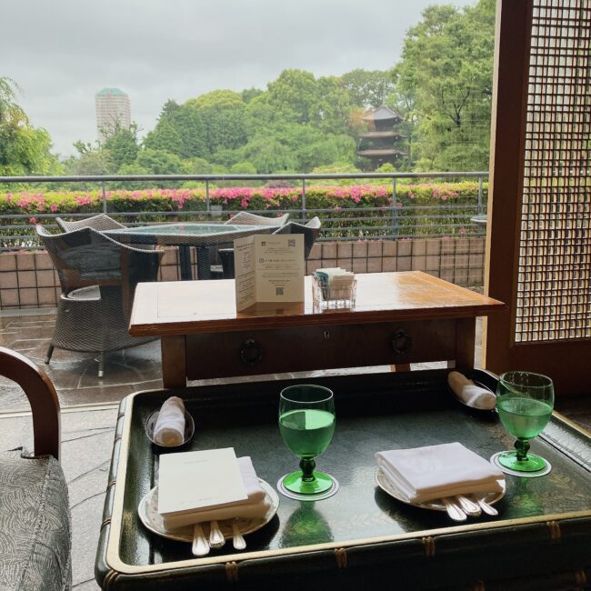 ホテル椿山荘東京「ル・ジャルダン」の窓際席から、緑豊かな庭園と歴史ある三重塔を望む光景。手前にはグラスやカトラリーが整えられたテーブルが写っており、奥には屋外テラス席と鮮やかなピンクのツツジが見える。自然光が差し込む開放的な空間。
