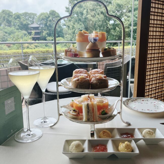 ホテル椿山荘東京「ル・ジャルダン」の円卓に置かれた三段のアフタヌーンティーセット。上段にはミニタルトやグラスデザート、中段にはスコーン、下段にはサンドイッチやセイボリーが美しく並べられている。手前にはシャンパングラス2脚と、ジャム、クロテッドクリームなどのコンディメントが白い小皿に盛られている。背景には緑豊かな庭園と、その奥に歴史ある三重塔が見える窓からの景色が広がっている。