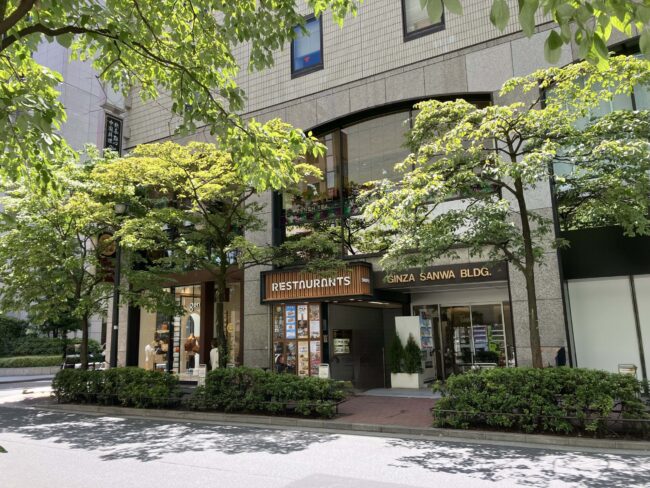 銀座松屋通り沿いに建つグレーのビル「GINZA SANWA BLDG.」の外観。1階には複数の飲食店が入っており、看板には「RESTAURANTS」の文字。右側の入り口の上には「GINZA SANWA BLDG.」と表示されている。緑豊かな街路樹が日差しを遮っている。マリアージュフレール銀座松屋通り店はこのビルの2階。
