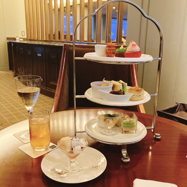 三段重ねのアフタヌーンティースタンドと飲み物、デザートが置かれたテーブル。上段にはカラフルなミニデザート、中段にはセイボリーとスコーン、下段にはセイボリーが並ぶ。落ち着いた雰囲気のラウンジでのアフタヌーンティーの様子。