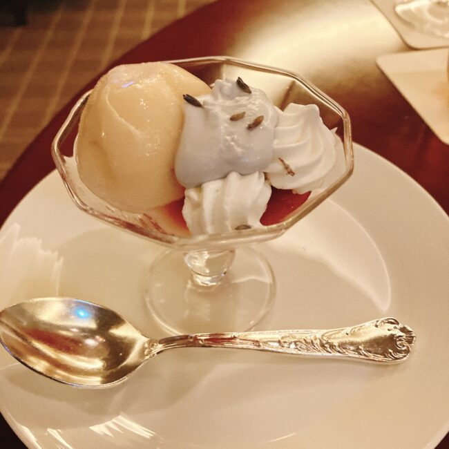 グラスに盛り付けられた、白桃のシャーベット、紫色のラベンダー風味のクリーム、白い生クリーム、赤いベリーのコンフィチュール。銀色のスプーンが添えられている。