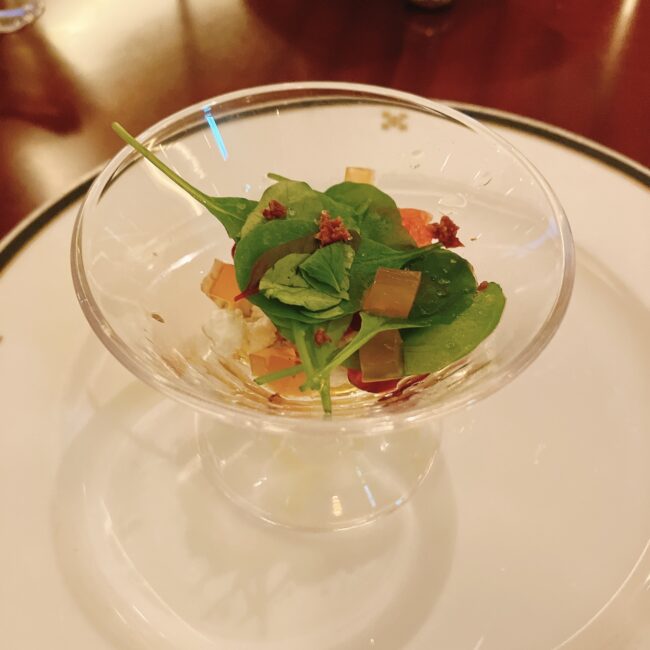 透明なステム付きの小さなグラスに入ったサラダ。ベビーリーフ、ピンクグレープフルーツの果肉、白いカッテージチーズ、透明なジュレ状のもの、赤いザクロの粒、カリカリに焼かれたベーコンが盛り付けられている。