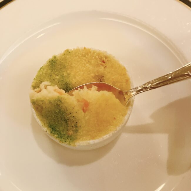 白い器に入ったフィッシュパイ。香草パン粉とチーズパン粉が乗った表面に、ゴールドのスプーンが差し込まれ、中身のクリーミーなクリーム煮が見えている。