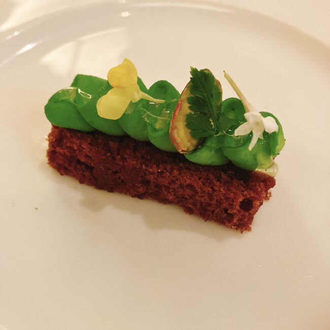 白い皿に盛られた、レモンドリズルケーキ。茶色いスポンジ生地の上に、緑色のクリームが絞られ、黄色い花と葉っぱが飾られている。
