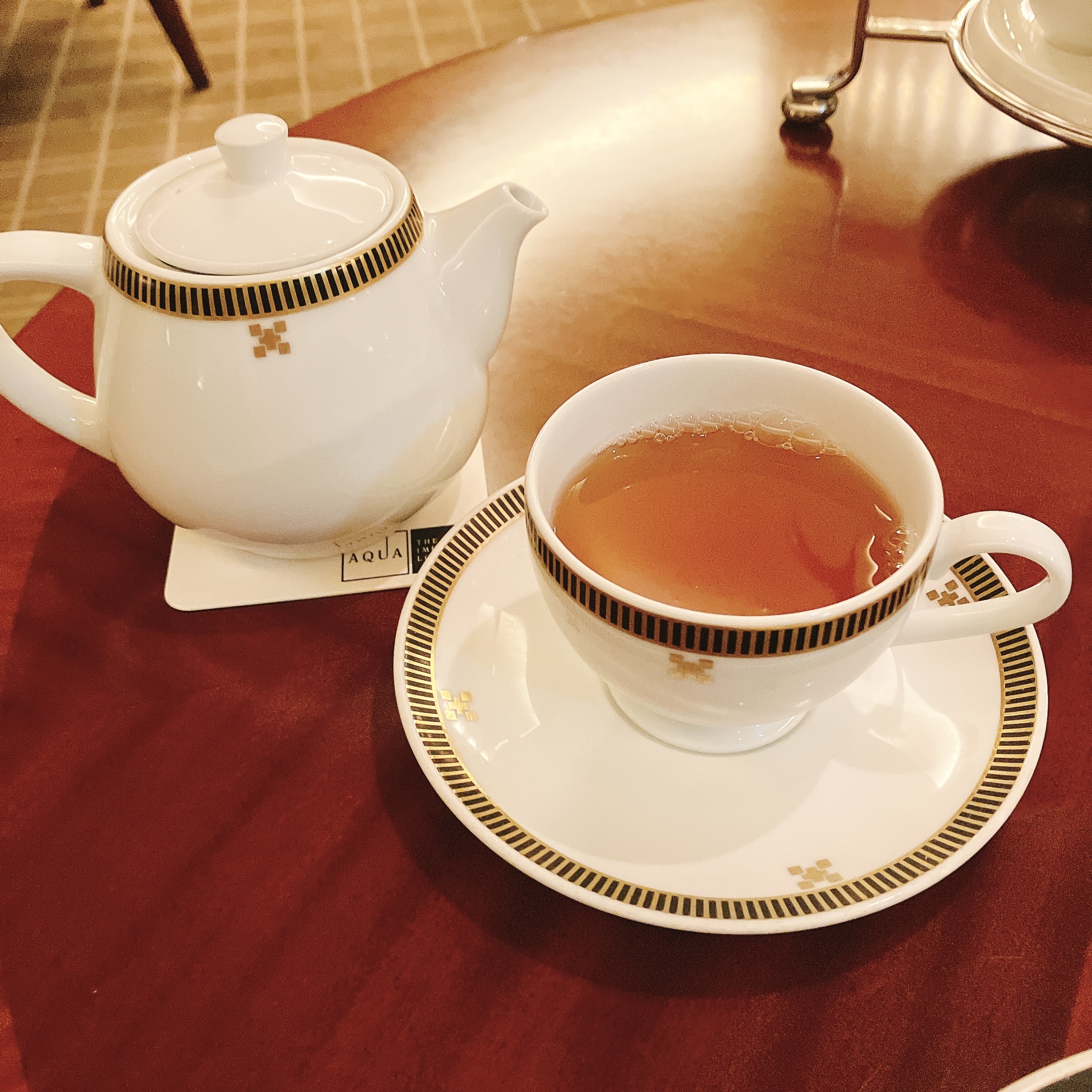 帝国ホテル 東京本館17階「インペリアルラウンジ アクア」のアフタヌーンティーで提供される、帝国ホテルのロゴマーク入りティーポットとティーカップ。カップには美しい琥珀色の紅茶が注がれており、木目の高級感のあるテーブルに置かれている様子。