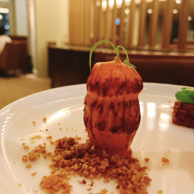人参の形をしたキャロットケーキが盛り付けられている。緑色の葉っぱが添えられ、ケーキの下には土をイメージしたクランブルが散らされている。