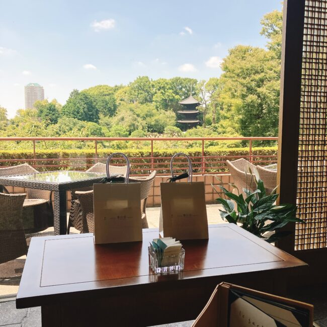 ホテル椿山荘東京のカフェ「ル・ジャルダン」の窓側のテラス席。目の前には、緑が生い茂る庭園と三重塔が見える。テーブルの上には、おみやげ用の紙袋が2つ置かれている。