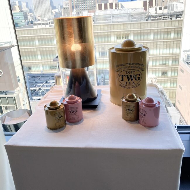 ザ・グラン銀座の窓辺のテーブル。白いテーブルクロスの上に、TWGの紅茶缶が大小合わせて5つ置かれている。背景には、明るい窓から見える高層ビル群の景色が広がっている。テーブルランプの柔らかな光が、紅茶缶を照らしている。