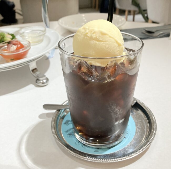 リチュエルカフェのコーヒーフロート。大きなグラスにたっぷりの氷とアイスコーヒーが注がれ、その上に大きなバニラアイスクリームが丸く乗っている。グラスは銀色のコースターに置かれている。背景には、アフタヌーンティースタンドと、小さなセイボリーが載った小皿が見える。