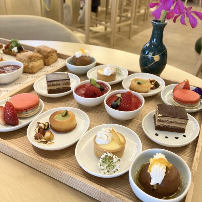 東京エディション銀座のアフタヌーンティー。木製のトレーの上に、様々な種類の小さくてカラフルなスイーツが白い小皿に一つずつ盛り付けられている。マカロン、タルト、ムース、ケーキなど、見た目も華やかで独創的なデザートが並ぶ。トレーの後ろには、スコーンとコンディメント、花瓶に入ったピンクの花が見える。
