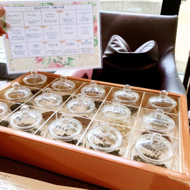 セルリアンタワー東急「坐忘」のアフタヌーンティー。木製の箱に入ったたくさんのガラス製の瓶があり、それぞれに様々な種類の紅茶やハーブティーの茶葉見本が入っている。瓶には「ダージリン」「ピーチメルバ」「カモミール」といった茶葉名が書かれた蓋が付いている。箱の後ろには、同じ茶葉名が記載された紙のメニューも見える。