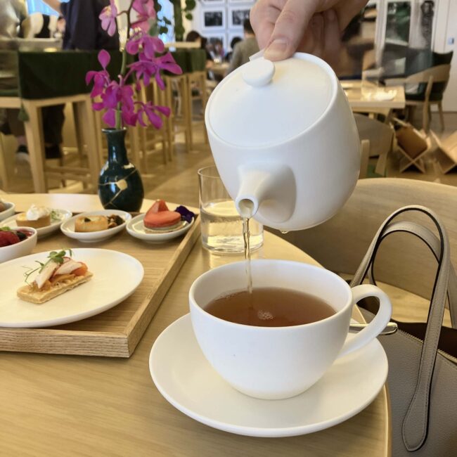 東京エディション銀座のテーブル。白いティーポットから、白いティーカップに琥珀色の紅茶が注がれている。テーブルの上には、木製のプレートに載った可愛らしいスイーツやセイボリー、そして水が入ったグラスが見える。背景には、モダンでおしゃれなカウンターの様子が写っている。