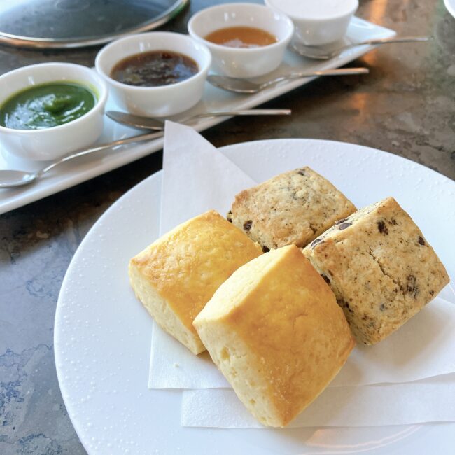 ジャヌ東京のアフタヌーンティーで提供されたスコーン。白い丸皿にプレーンスコーンとチョコレートチップ入りのスコーンが各2個ずつ、計4個盛り付けられている。奥には、江戸菜と柚子白餡、ジャム、糀みつ、豆乳ホイップなどの様々なコンディメントが並べられている。
