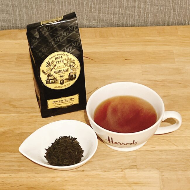 木目調のテーブルの上に、マリアージュフレール（Mariage Frères）の「プランス ドゥ コロンボ（PRINCE DE COLOMBO）」の黒いパッケージと茶葉の入った白い小皿、そして「Harrods」のロゴが入った白い大きなティーカップが置かれており、カップには美しい赤茶色の紅茶が淹れられている。