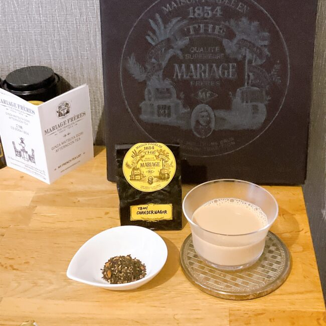 マリアージュ フレールの黒い本を背景に、スパイスが贅沢にブレンドされた茶葉「シャンデルナゴール」が白い小皿に盛られている。右側には、ガラスのカップに注がれた濃厚なロイヤルミルクティーがシルバーのコースターに置かれている。