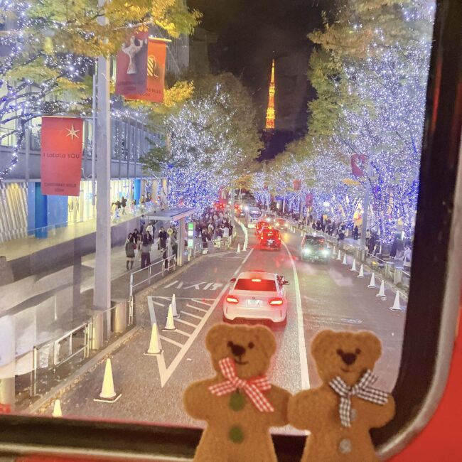 クリスマスシーズンにライトアップされた六本木けやき坂。街路樹のイルミネーションが輝き、車道には車、歩道には多くの人が行き交っている。遠くに東京タワーが見える。手前の窓枠には、ジンジャーブレッドマンのようなテディベアが2体並んでいる。