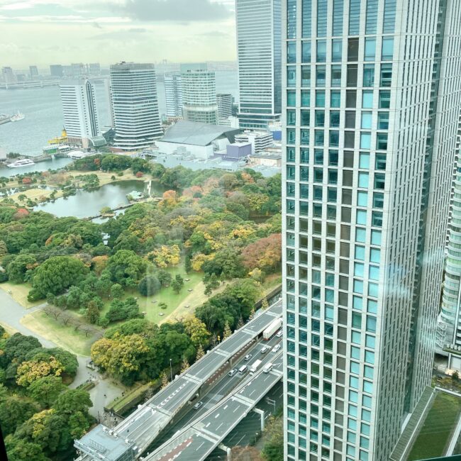 コンラッド東京のラウンジからの眺望。手前には高層ビルがそびえ立ち、その向こうに、緑豊かな浜離宮恩賜庭園の池と、遠くに広がる東京湾、そして高層ビル群が一望できる。空は曇り空で、都会的な景色の中に自然の緑が鮮やかに映えている。