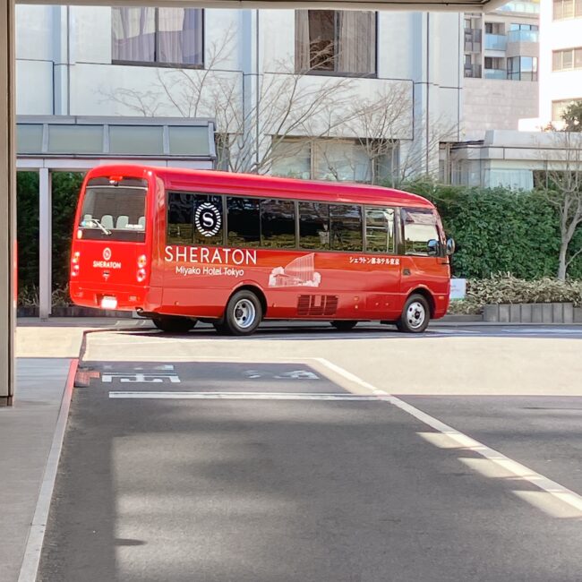 シェラトン都ホテル東京の正面外観。白い高層ビルと、ホテル名が書かれたエントランス。敷地内には植栽と、ホテルバスとみられる赤いバスが見える。	