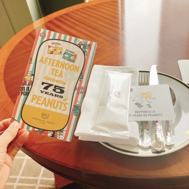 帝国ホテルのスヌーピー75周年記念アフタヌーンティーのテーブルセッティング。カトラリーとナプキン、そして「AFTERNOON TEA Celebrating 75 YEARS of PEANUTS」と書かれたメニューカード。