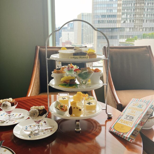 帝国ホテル東京「インペリアルラウンジ アクア」のスヌーピー75周年記念アフタヌーンティーの全体像。3段のティースタンドには、王冠スヌーピーパン、チャーリーブラウンバーガー、スヌーピーチーズケーキなどキャラクターモチーフのスイーツやセイボリーが並び、別添えのスペシャルスイーツも確認できる。窓の外には日比谷公園と高層ビルが広がる絶景。