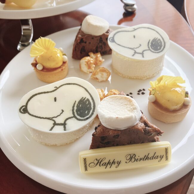 帝国ホテル スヌーピーアフタヌーンティーの3段目スイーツの全景。スヌーピーの顔が描かれたチーズケーキ、ブラウニー、タルトが盛り付けられ、手前には「Happy Birthday」のメッセージプレートがある。