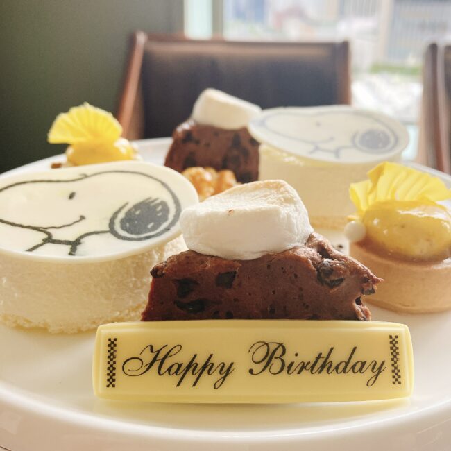 帝国ホテル東京のスヌーピーアフタヌーンティーのスイーツに、黄色のチョコレートプレート「Happy Birthday」が添えられた写真。スヌーピーの顔が描かれた白いケーキや、マシュマロが乗ったスコーンが確認できる。