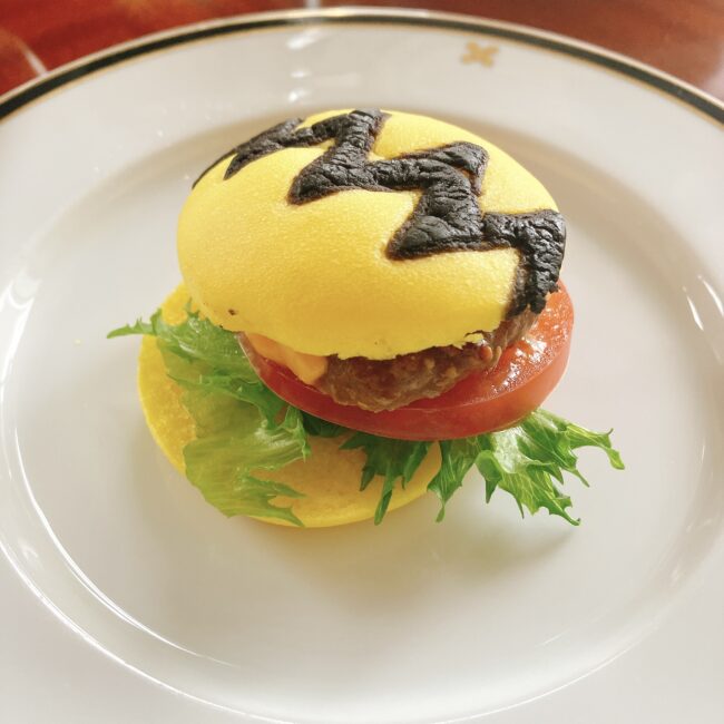 帝国ホテル スヌーピーアフタヌーンティーのBBQバーガーのクローズアップ。チャーリーブラウンのギザギザ模様の黄色いバンズと、ジューシーなパティ、トマト、レタスが挟まれたミニハンバーガー。