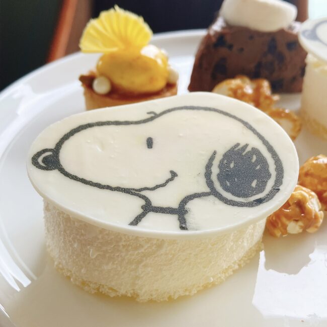 帝国ホテル スヌーピーアフタヌーンティーのメインスイーツ「スヌーピーのブルーベリーチーズケーキ」のクローズアップ。スヌーピーの顔が描かれたホワイトチョコが乗り、極上の口どけのチーズケーキの中にブルーベリーソースが入っている。