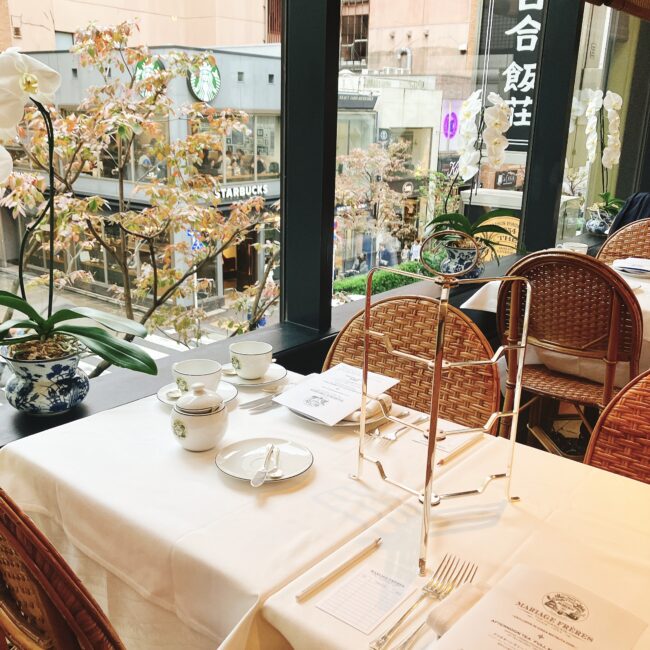 マリアージュフレール銀座松屋通り店の窓際席。窓の外には紅葉が始まった街路樹と銀座松屋通りが見える。テーブルには白いリネンとアフタヌーンティーの準備が整っている。