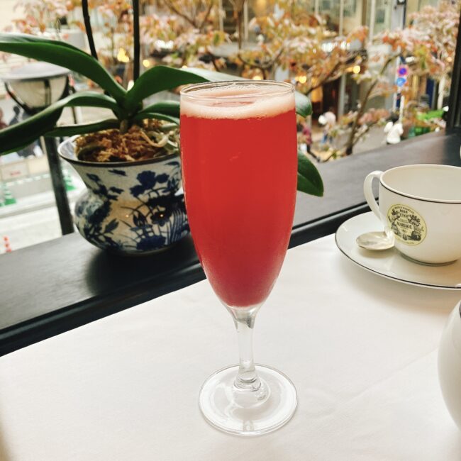 マリアージュフレール銀座松屋通り店の25年秋「フルムーンパーティーアフタヌーンティー」で提供された、赤色の「ルージュメティス」をベースにしたティーモクテル。窓の外には秋の街路樹が見える。