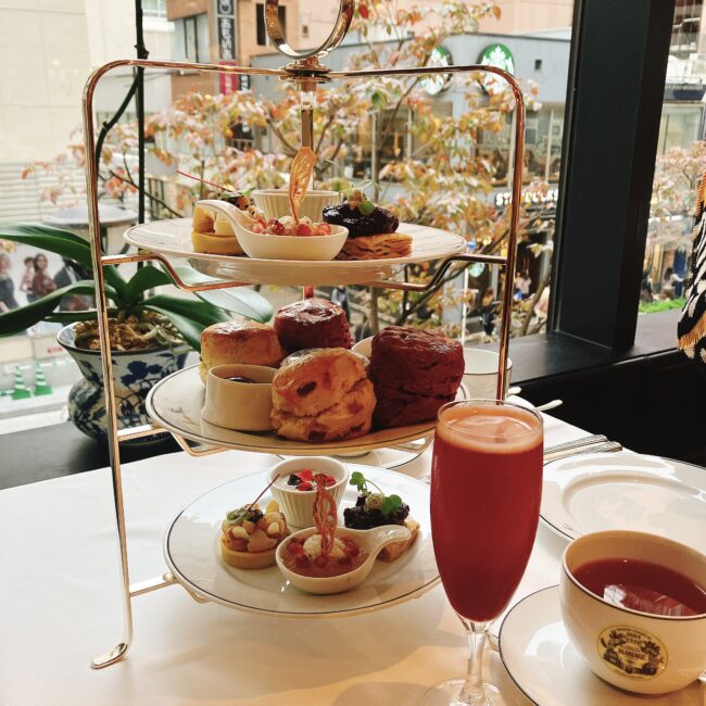マリアージュフレール銀座松屋通り店で提供された、2025年秋の「フルムーンパーティーアフタヌーンティー」の三段スタンド全体像。鮮やかな赤色を基調としたセイボリーとスコーンが盛り付けられ、手前にはティーモクテルと紅茶のカップが置かれている。背景には秋の街路樹が見える。