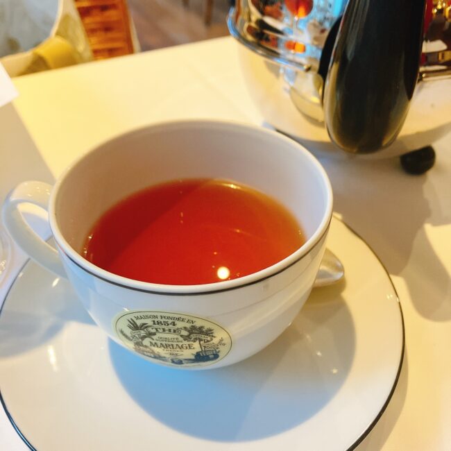 マリアージュフレールの白いティーカップに注がれた、艶やかな赤褐色の紅茶「プレーン ルヌ」。満月をテーマにした黒茶で、アーモンド、ハチミツ、希少なスパイスがブレンドされた甘く複雑な味わいが特徴。