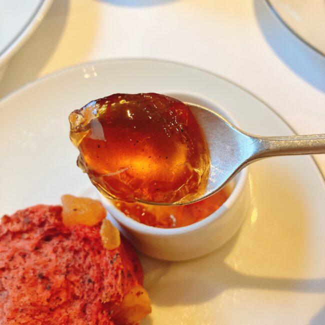 スプーンで掬い上げた、琥珀色に輝くマリアージュフレールの紅茶ジャム「マルコポーロ」。ジャムの中には茶葉の粒子が見え、手前には限定の赤色のスコーンの一部が写っている。