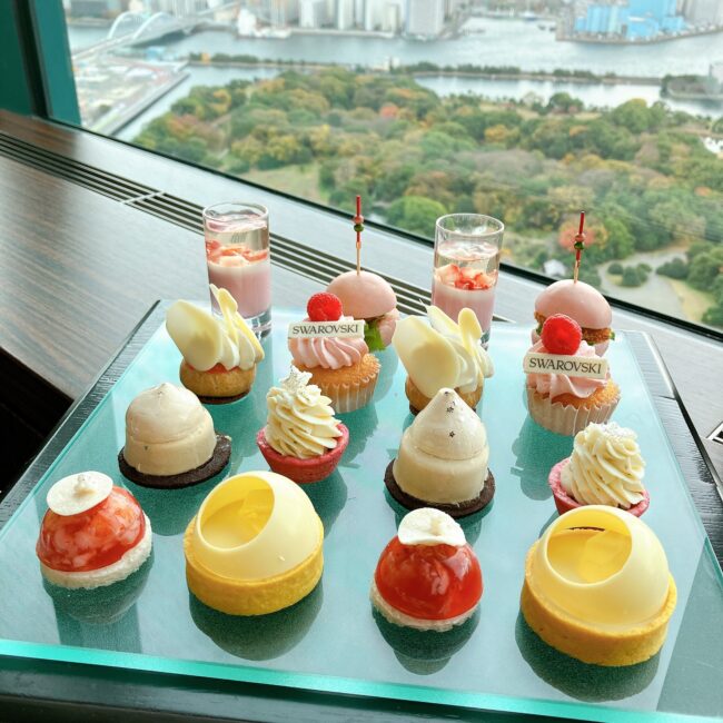 コンラッド東京のバー＆ラウンジ トゥエンティエイトで提供されているアフタヌーンティーのティーフーズ。ガラスのプレート上に赤、白、黄色のカラフルなスイーツと小さなハンバーガー状のセイボリーが均等に並べられ、背景の大きな窓からは東京湾と浜離宮恩賜庭園の絶景が見える。