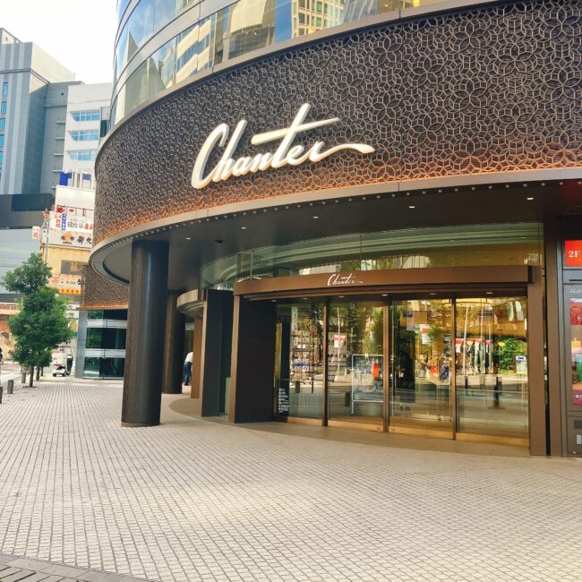 東京・日比谷にある商業施設「日比谷シャンテ」の外観。曲線を描くガラス張りの建物上部には「Chanter」のロゴが大きく掲げられ、シックな茶色の模様が施されている。歩道には緑の木々が植えられ、都会的で洗練された雰囲気。