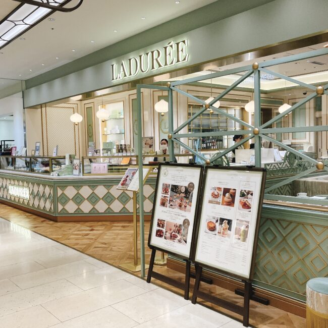 日比谷シャンテ1階にあるラデュレの店舗入り口。パステルグリーンのアイコニックな外観とゴールドの縁飾りが特徴で、「LADUREE」のロゴが輝いている。手前にはアフタヌーンティーや限定メニューの案内スタンドが二つ置かれている。