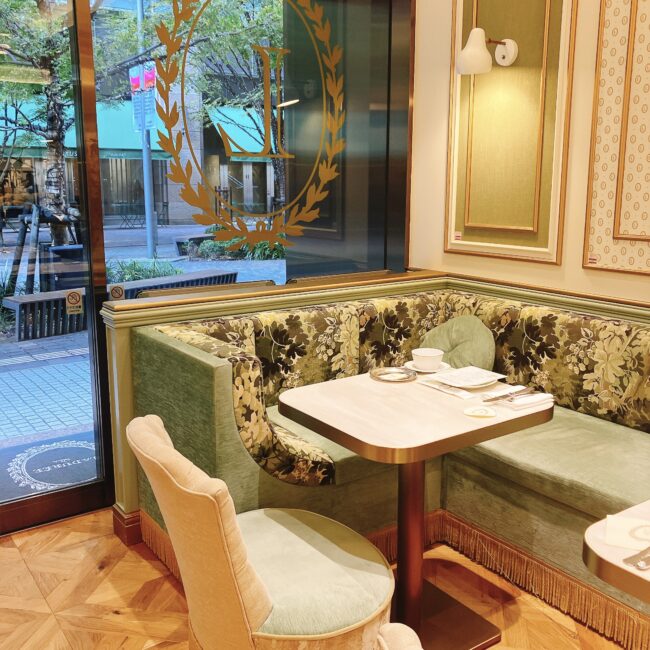 ラデュレ日比谷の窓側の角席。パステルグリーンとベージュを基調とした内装で、花柄のソファとピンクベージュの椅子、ゴールドフレームの小さなテーブルが配置されている。窓の外には日比谷中通りの街路樹が見え、一人でもくつろげる優雅な雰囲気。