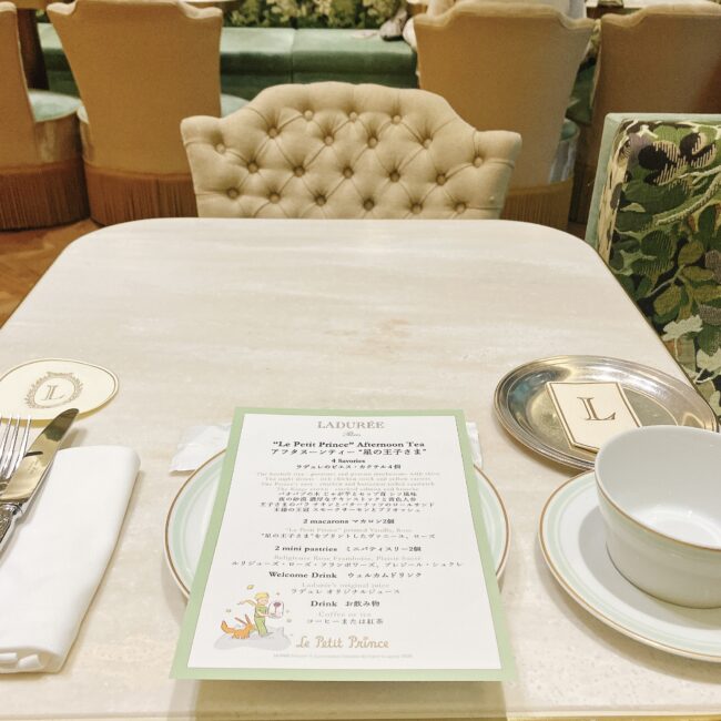 ラデュレ日比谷の席から見たテーブルセッティング。テーブルの上には、星の王子さまコラボのメニュー表、カトラリー、ナプキン、そしてラデュレオリジナルのティーカップとソーサーが置かれている。背景にはベージュとパステルグリーンを基調とした優雅なソファ席の背もたれが並ぶ。