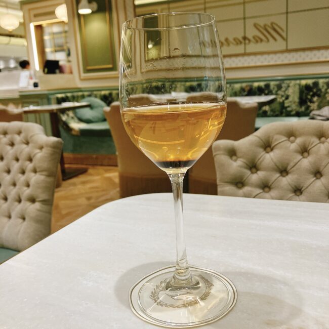 ラデュレ日比谷のアフタヌーンティーで提供された、ウェルカムドリンクのティーソーダ。ワイングラスに注がれた黄金色のドリンクは、「テ ジャルダン ブルー ロワイヤル」の紅茶をベースにしている。背景にはラデュレらしいパステルグリーンとゴールドの優雅な内装が見える。