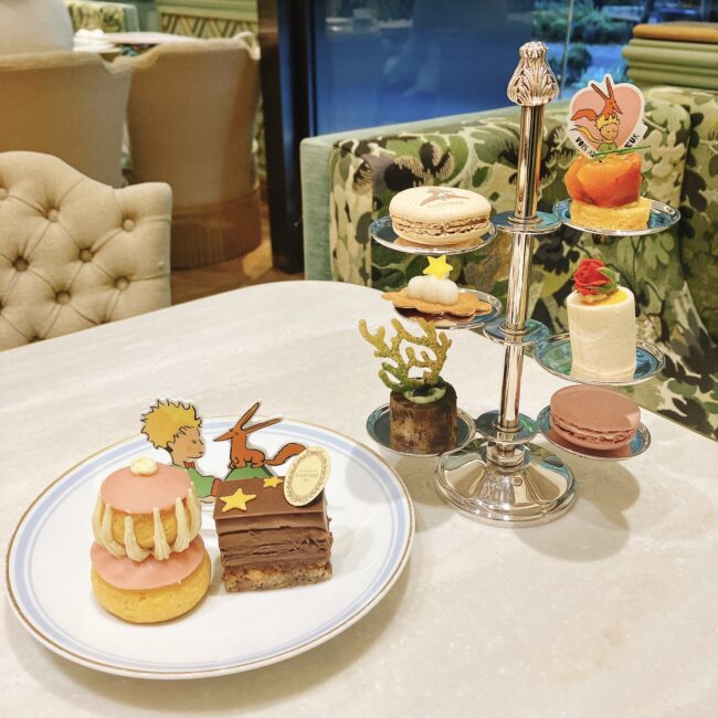 ラデュレ日比谷『星の王子さま』コラボのアフタヌーンティーのおひとり様用セット。銀色のスタンドにはマカロン、タルト、ミニパティスリーが並び、手前の白い皿には星の王子さまとキツネのプレートがついた2種のケーキが置かれている。背景は緑の花柄ソファの店内。