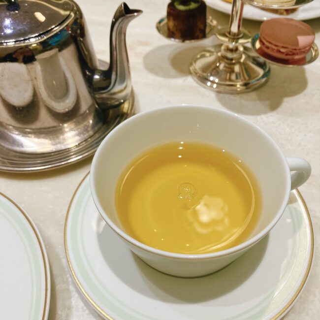 ラデュレ日比谷のアフタヌーンティーで提供された紅茶「テ ロワ ソレイユ」。カップには黄金色に輝く紅茶が注がれ、銀色のティーポットが隣に置かれている。中国の緑茶をベースに、キャラメル、ルバーブ、ベルガモットの香りが特徴。