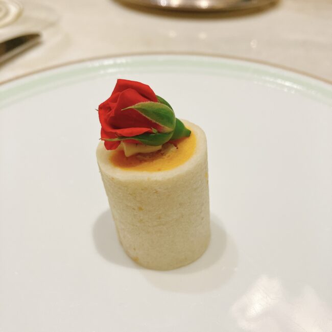 ラデュレ日比谷『星の王子さま』コラボアフタヌーンティーのセイボリー「王子さまのバラ チキンとバターナッツのロールサンド」。円筒形のサンドの上部には鮮やかな赤いバラの蕾と緑の葉が飾られ、バターナッツの黄色いクリームが覗いている。