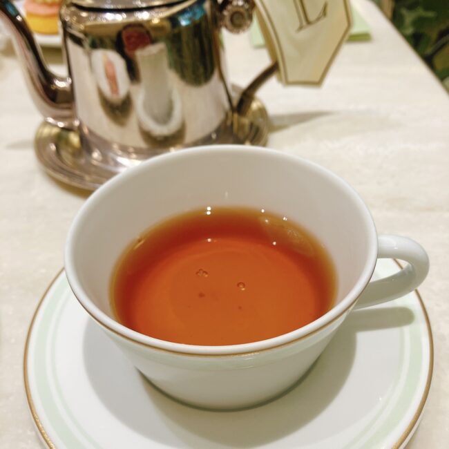 ラデュレ日比谷のアフタヌーンティーで提供された紅茶「テ マリー・アントワネット」。カップには琥珀色に輝く紅茶が注がれ、背景には銀色のティーポットが見える。中国紅茶をベースにバラ、ジャスミン、柑橘はちみつの香り、ドライフルーツの味が特徴。