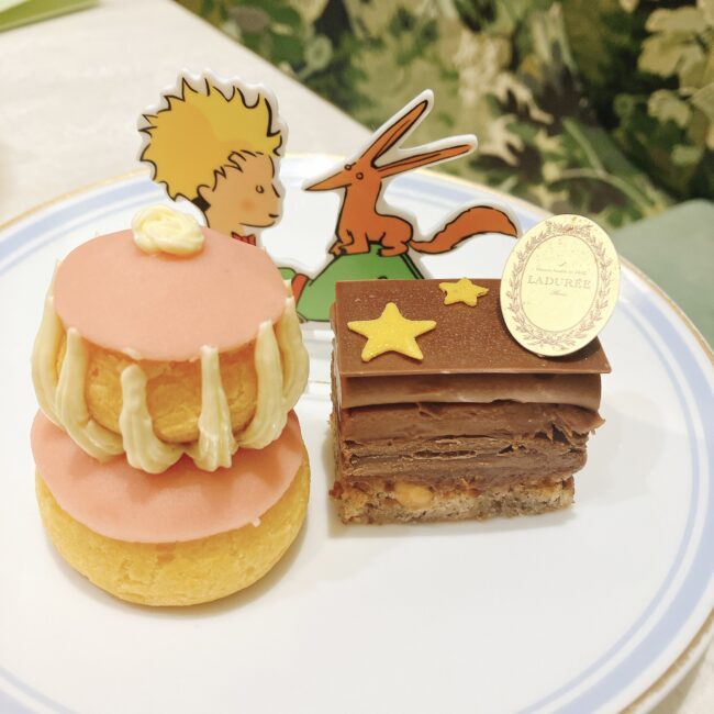 ラデュレ日比谷『星の王子さま』コラボアフタヌーンティーのミニパティスリー2種。左はピンク色のルリジューズ・ローズ・フランボワーズ、右は星の飾りとラデュレのプレートが乗ったチョコレートベースのプレジール・シュクレ。背景には星の王子さまとキツネのイラストのアクリルスタンドが添えられている。